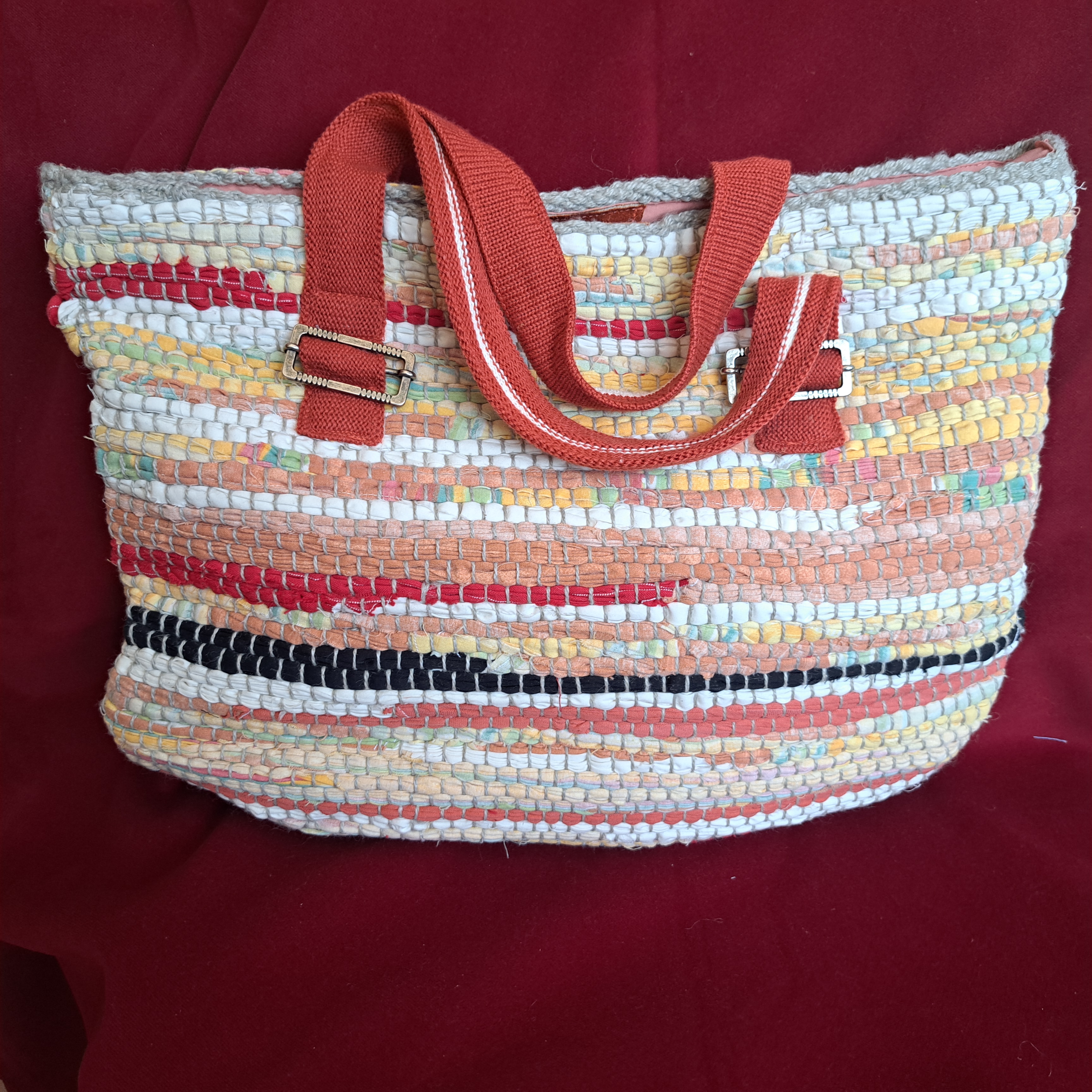 Handgefertigte Tasche aus recyceltem Stoff mit bunten Streifen, orangefarbenen Griffen und Metallschnallen, hergestellt im Handswork-Labor
