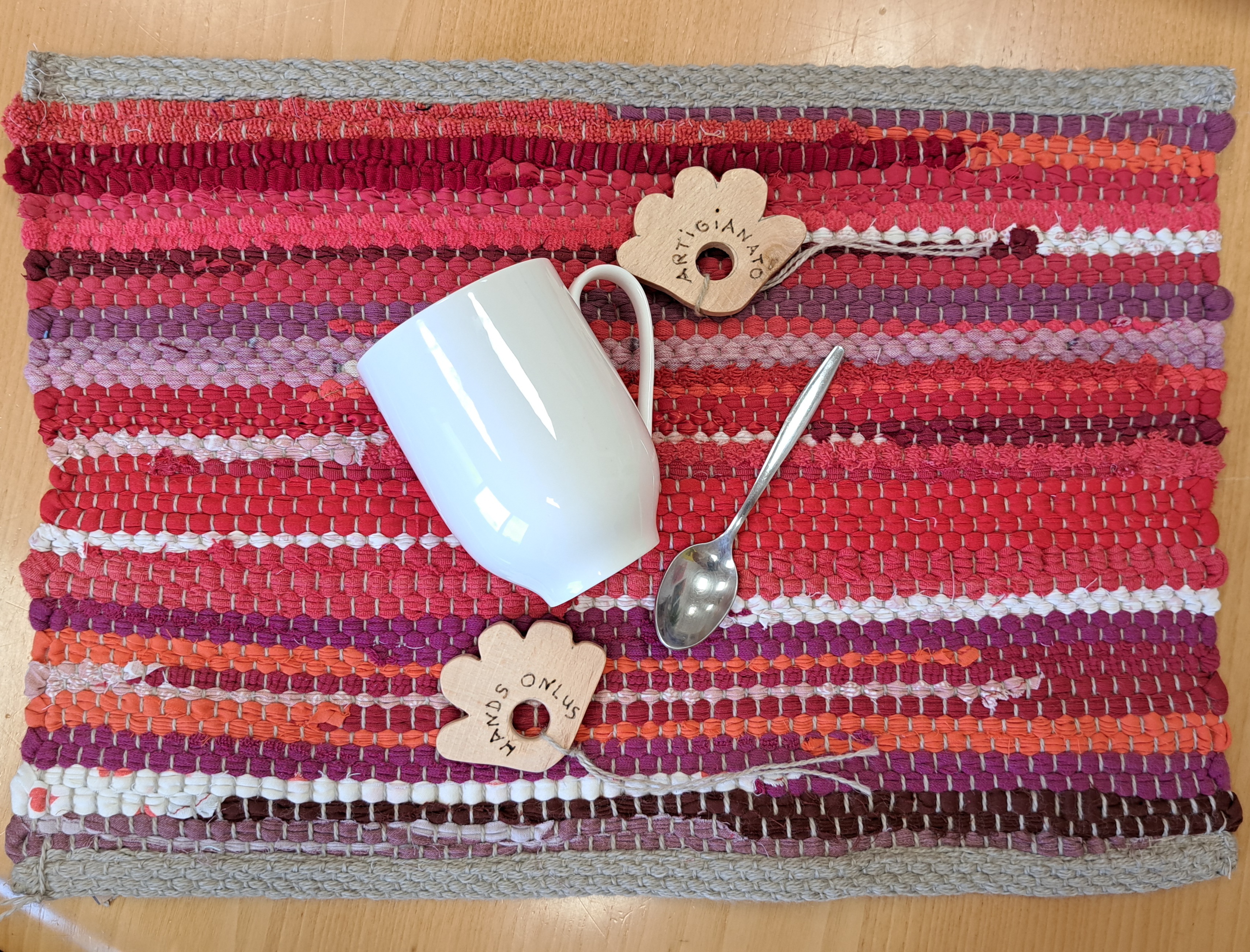 Handgemachtes Tischset aus rotem und lila recyceltem Stoff, mit einer weißen Tasse, Teelöffel und den Etiketten ARTIGIANATO und HANDS ONLUS
